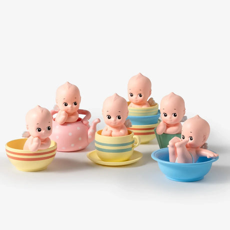 Six small baby doll figurines with various facial expressions sitting inside colorful tea cups, bowls, and a pink polka-dot teapot