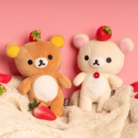Two Rilakkuma plush toys, including a cream-colored Korilakkuma Small Plush (20cm), sit on a beige blanket with strawberries scattered around them and a pink background, highlighting their cute look and premium softness.