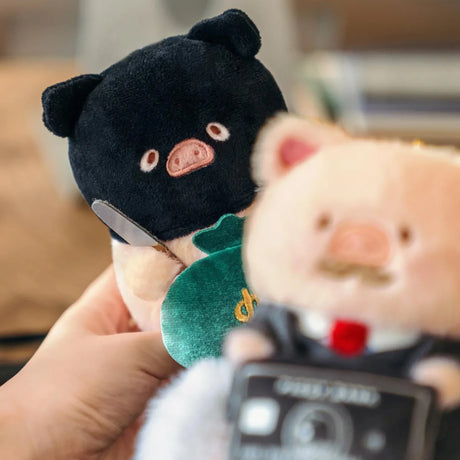 Black plush piggy keychain in green shirt behind a white piggy keychain.