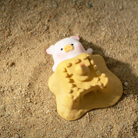 LuLu the Piggy figurine partially hidden under a yellow sandcastle mold on textured sandy beach surface