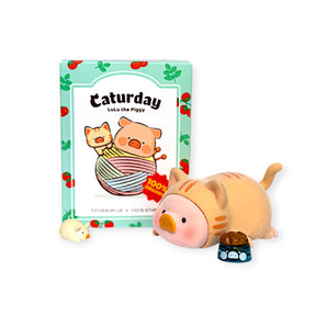LuLu the Piggy figurine dressed as a cat lying next to a food bowl and small mouse toy with Caturday box behind