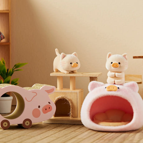 Two LuLu the Piggy figurines dressed as cats playing near a pig-shaped toy car and plush pet bed on wooden floor
