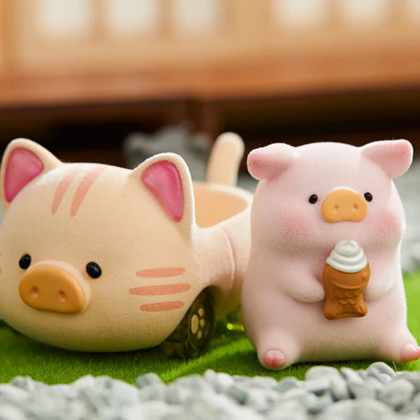 LuLu the Piggy figurine dressed as a cat sitting beside a pig-shaped toy car on green grass with small stones nearby