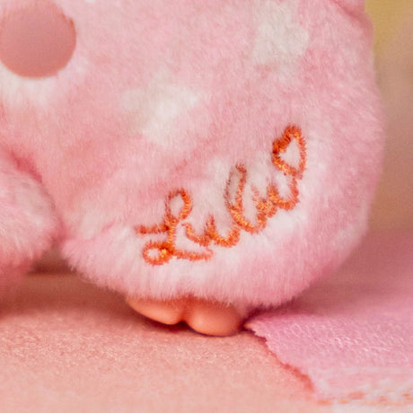 Close-up of pink plush fabric with embroidered "Lulu" signature and tiny plush feet on soft textured surface
