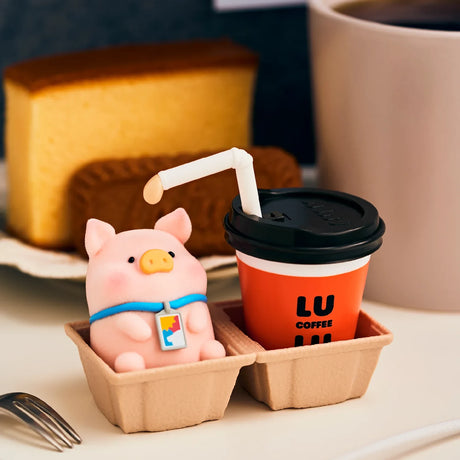 Piggy blind box figure seated in a small cardboard tray beside a coffee cup