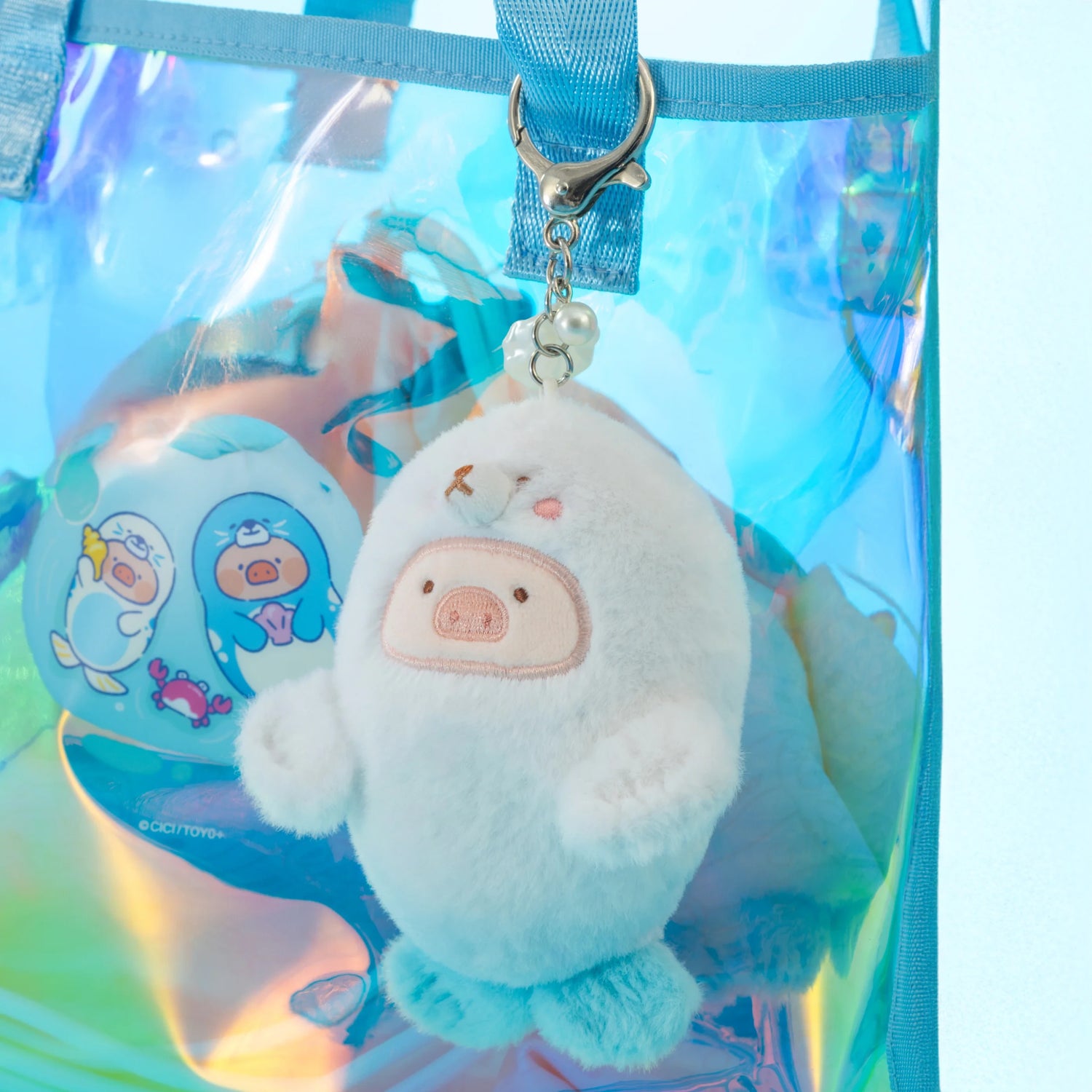 The LuLu The Piggy - Baby Seal LuLu Plush Keychain, shaped like a cute white and pink animal, hangs from a blue strap on an iridescent bag with cartoon designs—perfect for your next ocean adventure.