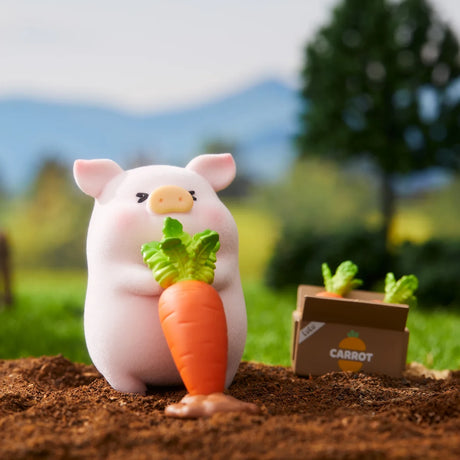 LuLu the Piggy figurine holding a large carrot in garden soil with a cardboard box labeled carrot in the background outdoors