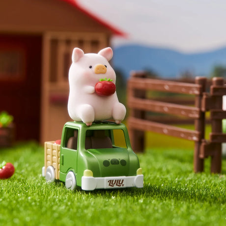 LuLu the Piggy figurine holding a red tomato sitting on top of a green toy truck on grass with farm background