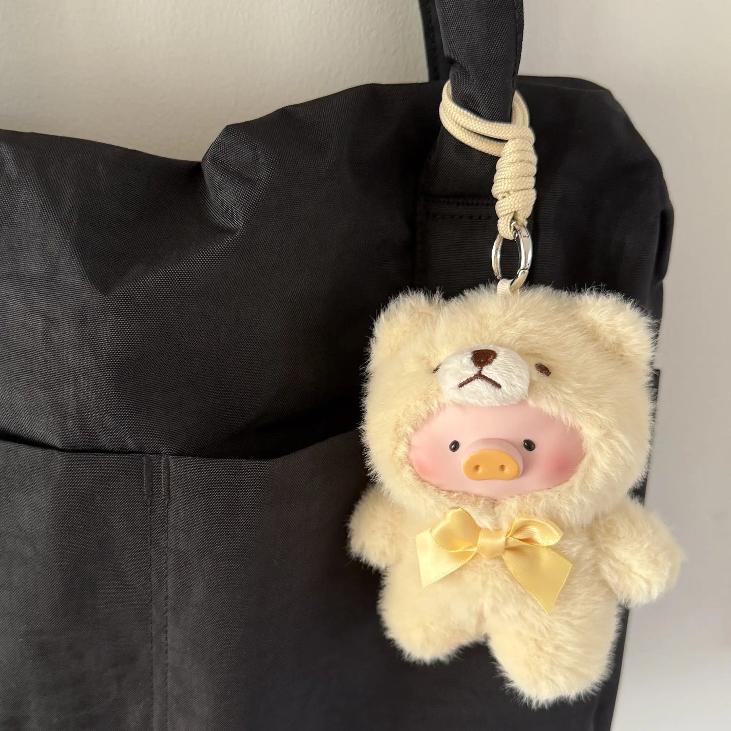 A Teddy LuLu the Piggy Plush Pal keychain by LuLu The Piggy dangles from the handle of a black fabric bag, featuring Teddy LuLu in a bear costume with a pig snout.