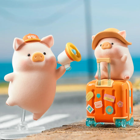 Two LuLu the Piggy figures, one with a megaphone and hat, the other sitting on a suitcase decorated with travel stickers by the sea