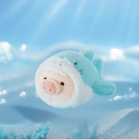 The LuLu The Piggy - Vibrating Shark Keychain features LuLu the Piggy with its face inside a blue shark costume, resting on a soft, light blue surface against a blurred, watery backdrop.