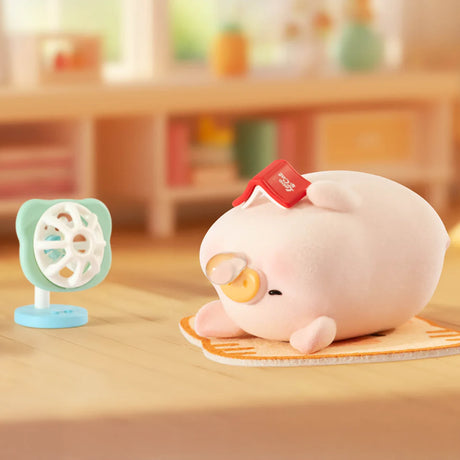 A LuLu The Piggy - Stay With You Blind Box figure by LuLu The Piggy rests on a mat with a red book on its head, facing a small blue fan in a softly lit, cozy room.
