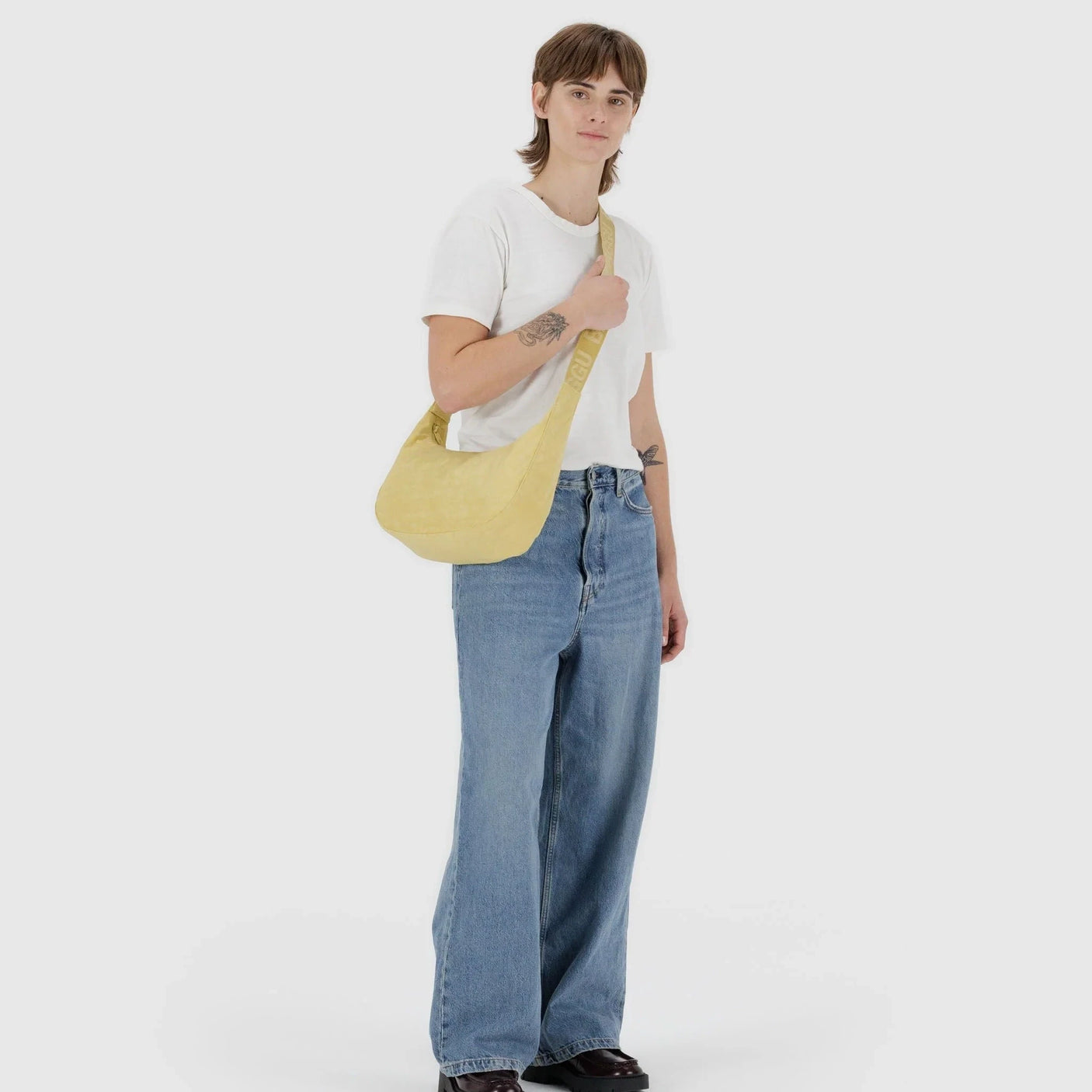 A person in a white t-shirt, blue jeans, and black shoes stands against a plain background carrying a Baggu Medium Nylon Crescent Bag - Butter. The bag's bright shade is truly sunshine ready.