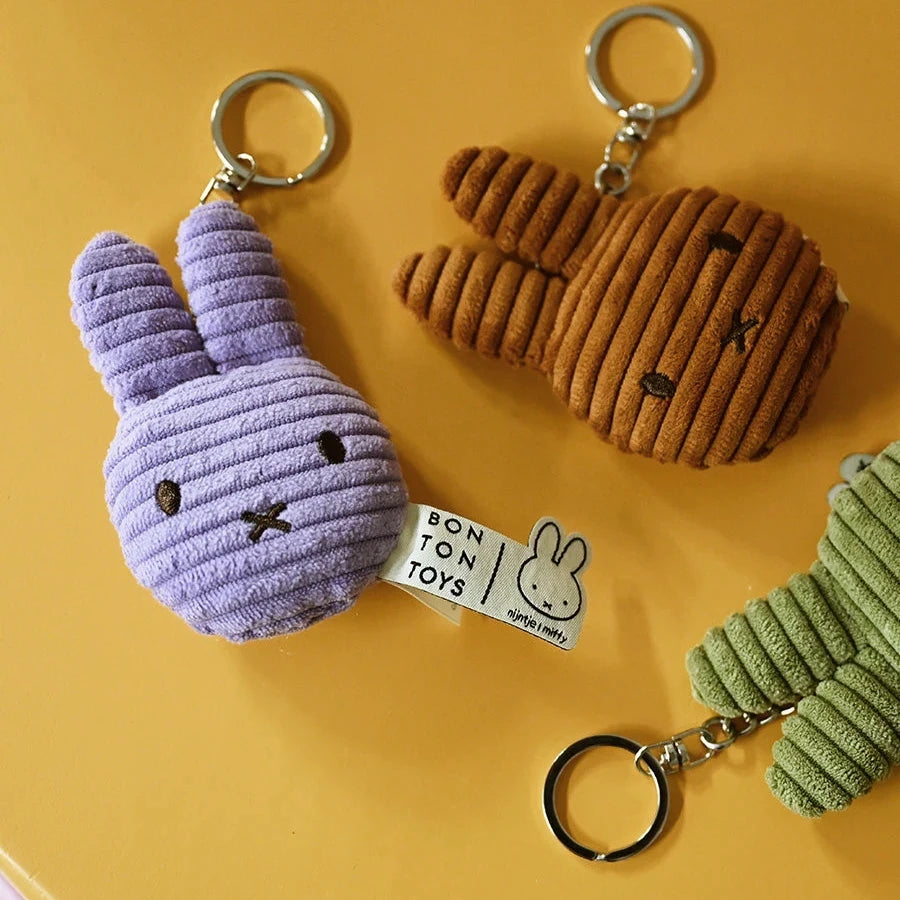 A yellow surface displays three Miffy keychains: the purple "Miffy Flat Corduroy Keychain" and two others, in brown and green, all crafted from recycled polyester.