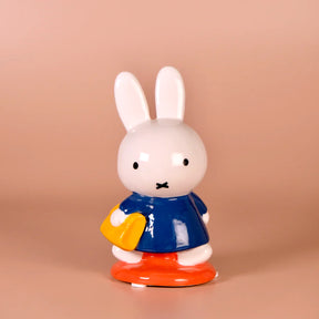 Glossy white ceramic bunny figurine wearing blue dress holding yellow purse on peach background
