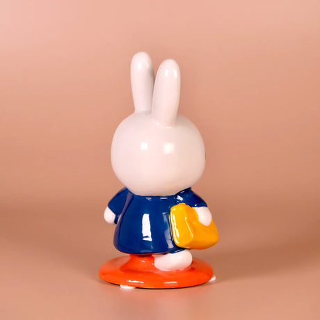Glossy ceramic bunny figurine in blue coat holding yellow bag standing on orange base against peach background