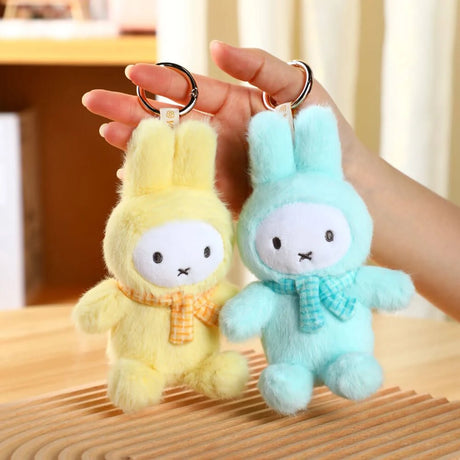 Hand holding pastel yellow and blue Miffy plush keychains with checkered scarves over wooden surface