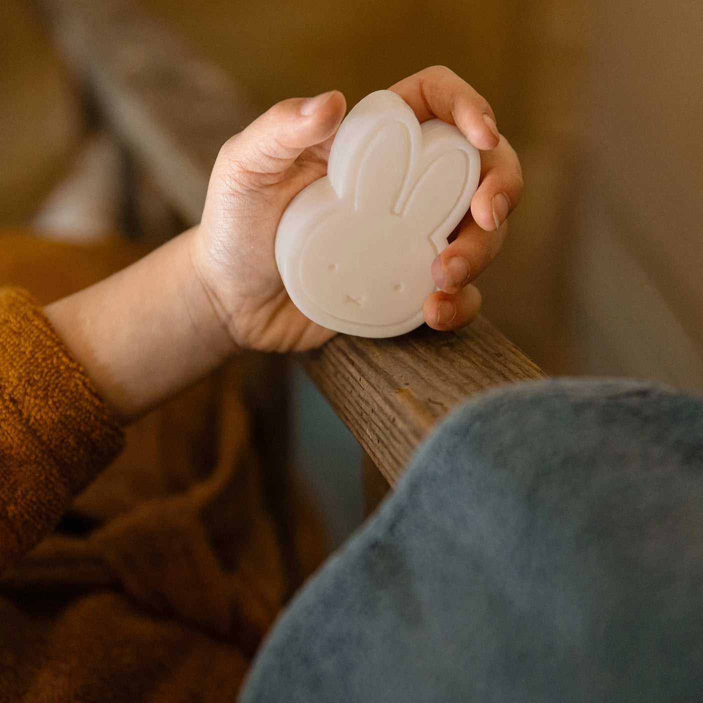 A child's hand clasps the Miffy Soap - Berry Parfum, a white bunny-shaped piece by Miffy, against a wooden railing. This vegan-friendly soap or toy embodies childhood's simplicity and joy while promoting a plastic-free future.