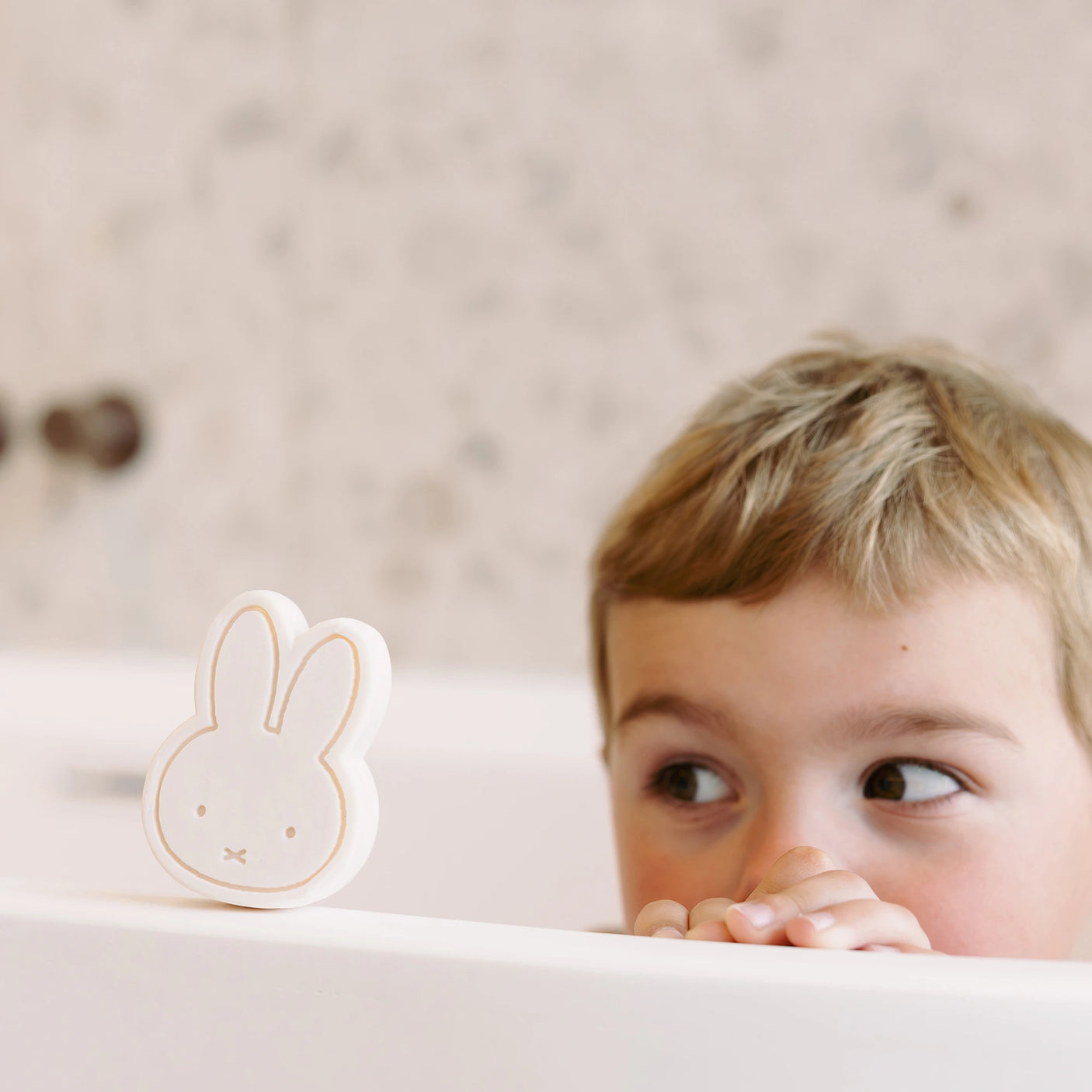 A young child peeks over the edge of a bathtub, eyes wide with wonder at a Miffy Soap - Berry Parfum shaped like a bunny, crafted to be vegan-friendly and plastic-free, against a blurred bathroom background.