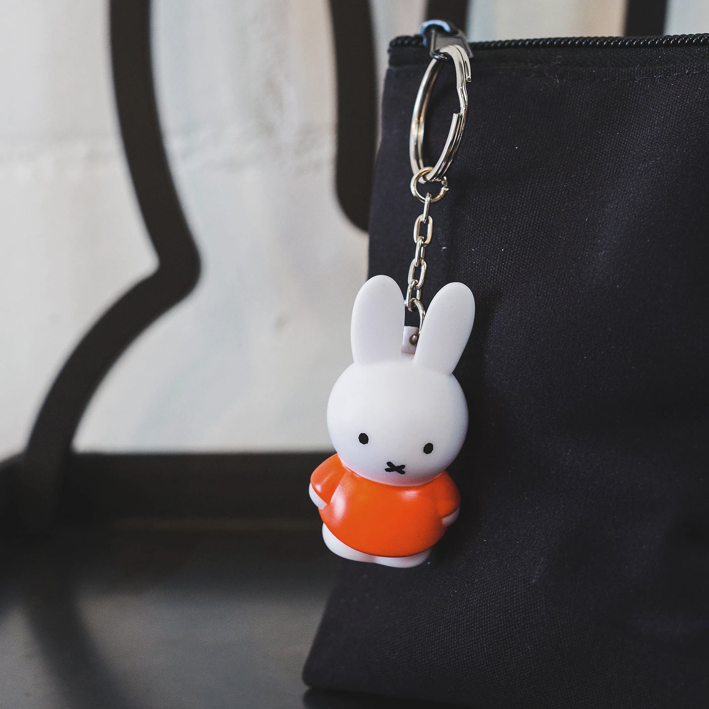 This charming Miffy Keychain - Classic from the Miffy brand showcases the iconic white rabbit character in an orange shirt, making it a perfect match for the timeless elegance of a sleek black bag.