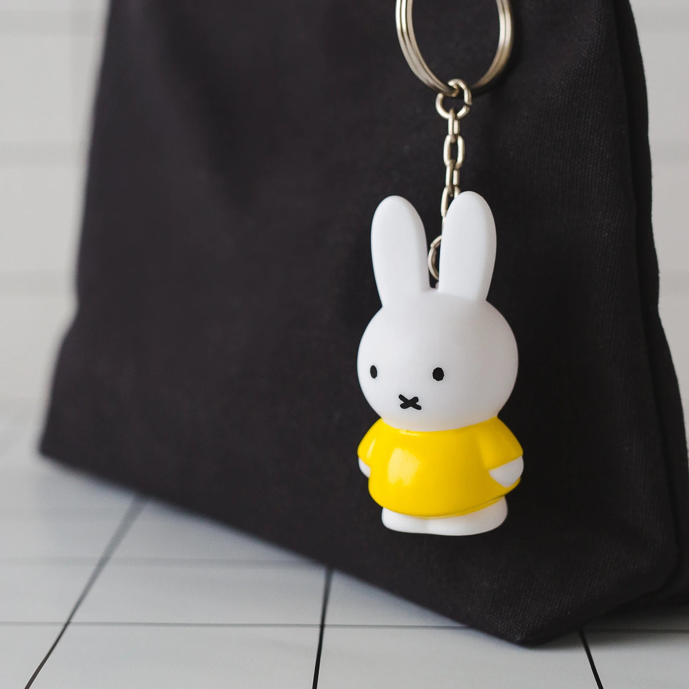 The Miffy Keychain - Classic by Miffy, featuring a charming design with a yellow shirt, adds a touch of classic colors as it dangles from a black bag on the tiled surface.