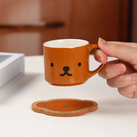 A hand holds a small brown Miffy 70th Bday Collectible Mug with a bear face, next to the matching coaster on a white surface—ideal for Miffy fans and lovers of unique blind box collectibles.