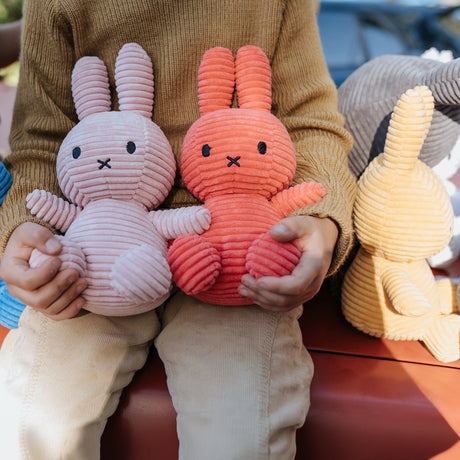 Seated with a Miffy Bunny Plush in each hand—one pink, one red—a person enjoys the company of these hypoallergenic companions. A Miffy Bunny Plush Corduroy in buttercream, crafted from recycled polyester, rests peacefully beside them.