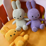 Three eco-friendly Miffy Bunny Plush Corduroy toys in gray, lilac, and yellow sit on a chair with multiple colors.