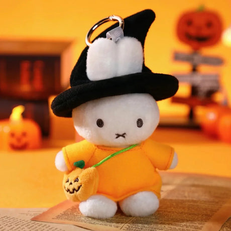 Miffy plush keychain in witch costume holding pumpkin bag with Halloween decorations blurred in background