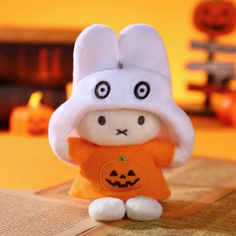 Plush Miffy keychain wearing a white ghost hood and orange shirt with jack-o'-lantern design on warm Halloween backdrop