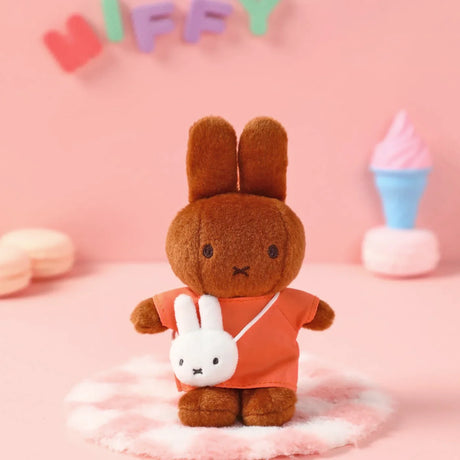 Brown plush bunny wearing orange outfit with small white bunny-shaped crossbody bag against pink backdrop