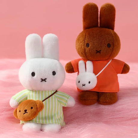 Two plush bunnies, one white in striped green outfit with brown bear bag, one brown in orange shirt with white bunny pouch on pink surface