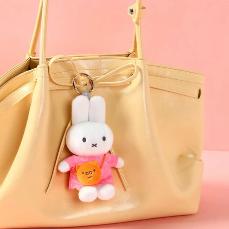 White plush bunny keychain wearing pink outfit with orange bear-shaped pouch hanging from beige handbag against pink background