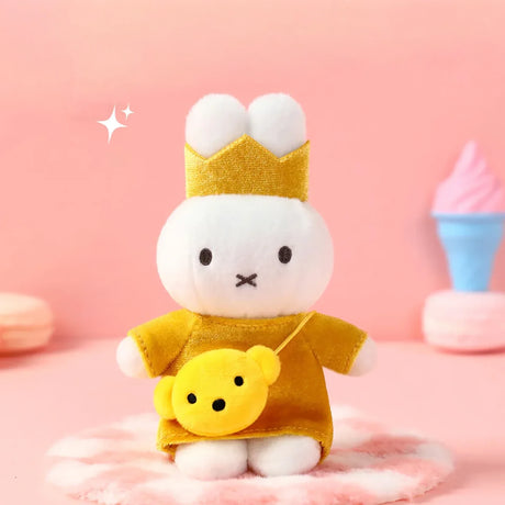 White plush bunny wearing gold crown and dress with yellow bear-shaped crossbody bag on pink fuzzy surface