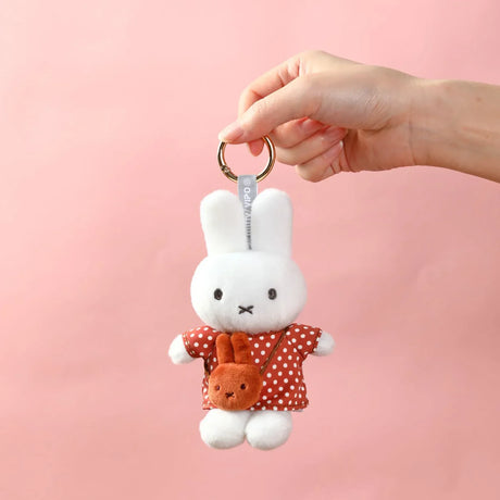 Hand holding white plush bunny keychain wearing red polka dot dress with small brown bunny purse on pink background