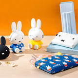 A Miffy Bunny Zipper Pouch 19cm in Blue, along with two Miffy rabbit figurines in blue and yellow, a white wallet, coins, a book, and blue folders sit on a table against an orange background. The pouch from Miffy adds versatile storage charm to this delightful setup.