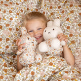 A child in a Vintage Flower Dress lies on a floral bedspread, smiling with delight while holding two Miffy Plush White & Vintage Flowers toys from the Miffy brand.