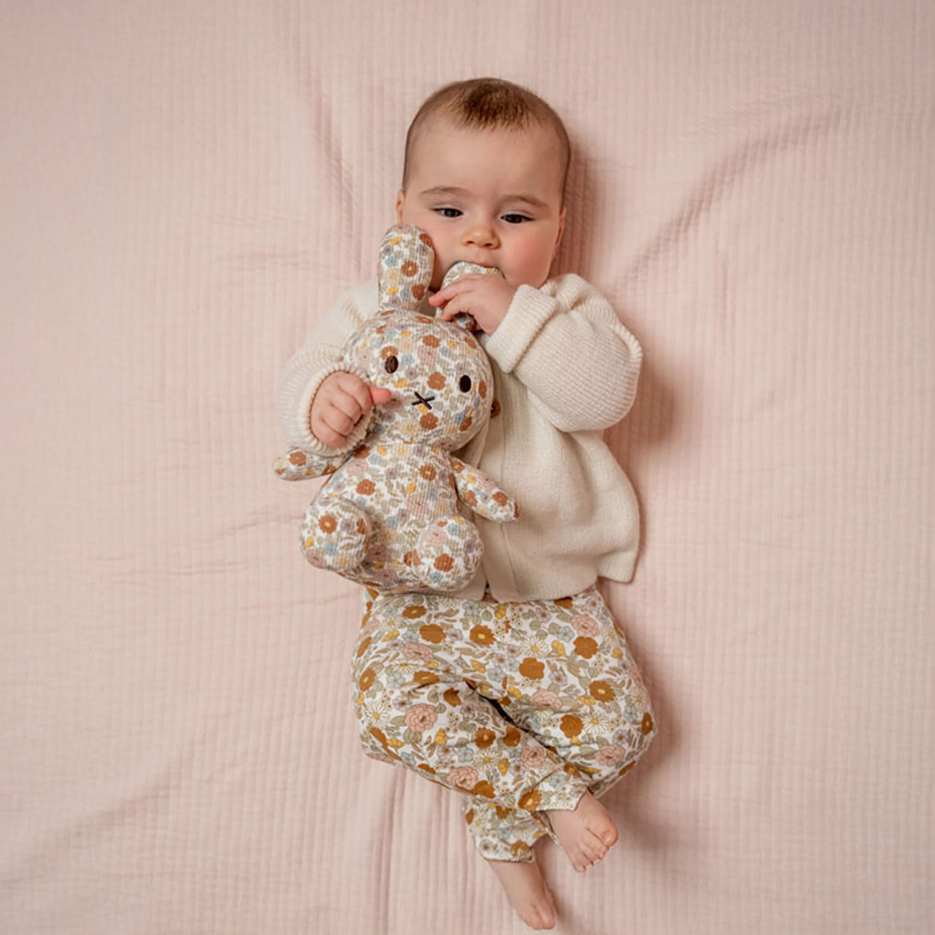 A baby lies on a pink blanket holding a 25cm Miffy Plush adorned with vintage flowers. They are wearing a white sweater and floral pants.