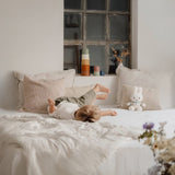 A child in casual clothes lies on a bed with pillows, accompanied by the Miffy Plush White & Vintage Stripes - 25cm, and stacked cups, basking in the soft daylight filtering through the window.