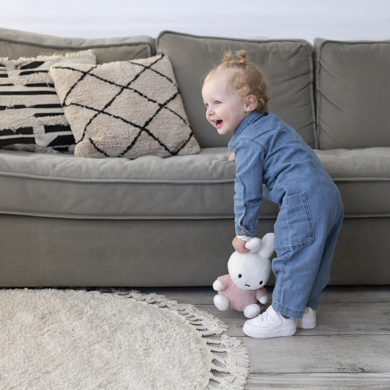 A toddler in a denim jumpsuit stands by a gray couch, holding the Miffy Plush Fluffy Plush Pink - 25cm from Miffy. The couch is adorned with patterned pillows, and a fluffy pink round rug adds charm to the nursery decor.