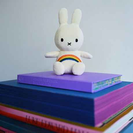 The 18cm Miffy Rainbow Plush, known for its super-soft texture and embroidered rainbow details, rests on a purple book atop a stack of colorful books.