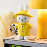 White plush bunny wearing yellow raincoat and hat standing on stacked books beside yellow lamp and zebra-striped mug