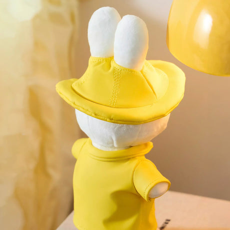 Back view of white plush bunny wearing yellow rain hat and matching yellow raincoat on table near yellow lamp