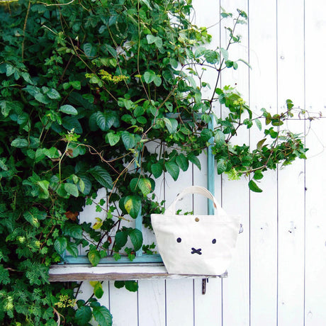 A Miffy Small Canvas Tote Bag, featuring a classic cotton canvas and simple face design, rests on a window ledge surrounded by leafy vines and a white wooden wall—an everyday essential for those who appreciate subtle elegance.