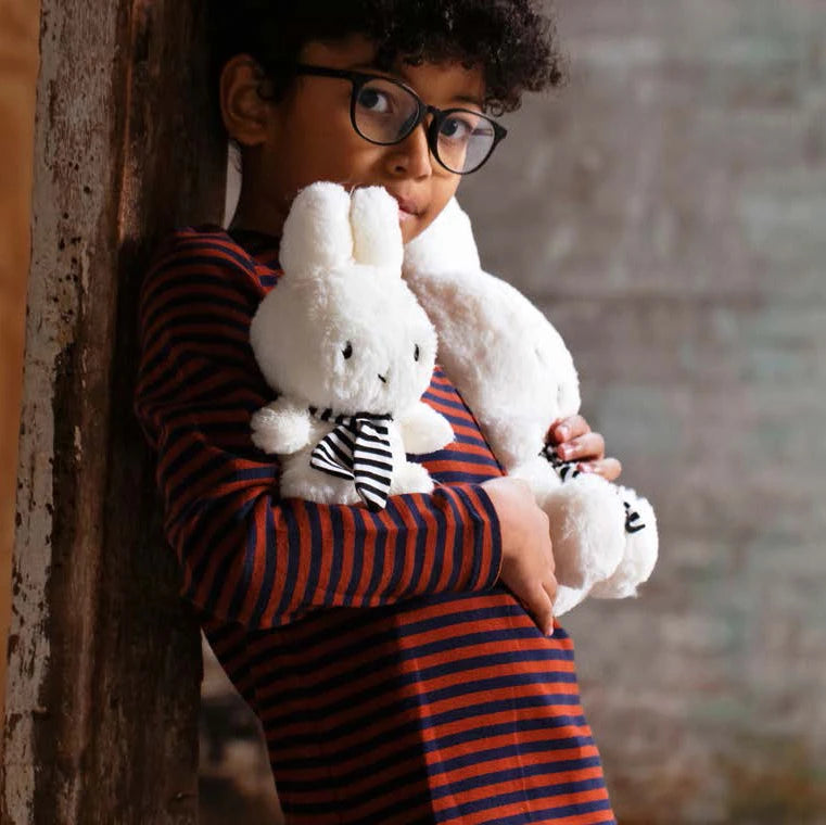 A child wearing glasses leans against a wooden surface, hugging two cuddly stuffed animals. One of them is the charming Miffy Bunny Plush with Scarf from the Miffy brand, crafted with recycled PET filling for enhanced softness and sustainability.