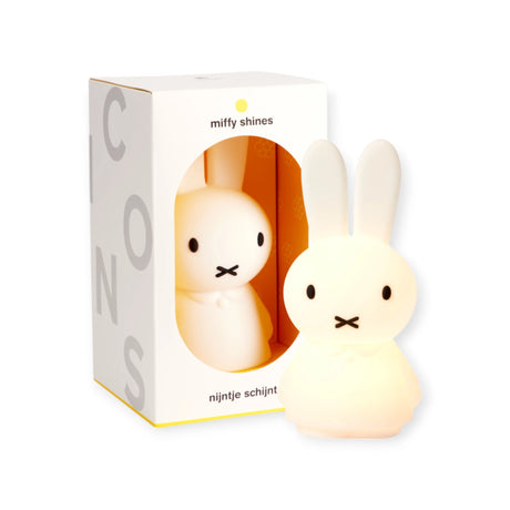 The Miffy Shines - Night Light (22cm), a charming rabbit-shaped lamp in white, sits beside its packaging box. This delightful nightlight from Miffy features touch dimming and is rechargeable, making it perfect for adding a gentle glow to any room.