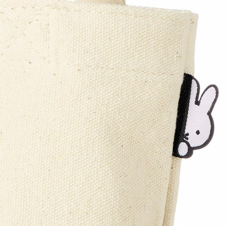 The Miffy Small Canvas Tote Bag features beige cotton canvas with a charming black and white bunny tag peeking from the side seam.