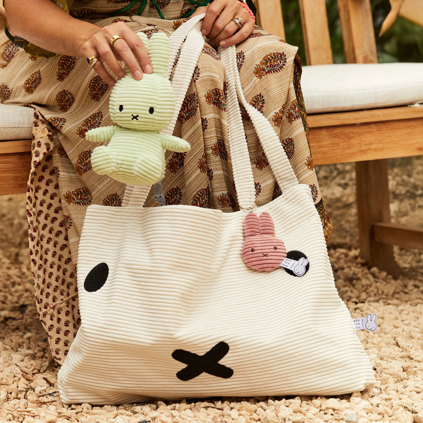 A person on a bench holds a plush green rabbit over an eco-friendly textured bag with a black 'X' design, featuring an off-white Miffy Flat Corduroy Keychain.