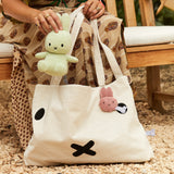 A person holds a green plush toy while seated on a wooden bench. Nearby, a large cream bag with an attached Miffy Bunny Plush Corduroy in lilac rests beside them. The setting is outdoors, surrounded by gravel and greenery, creating an eco-friendly ambiance.