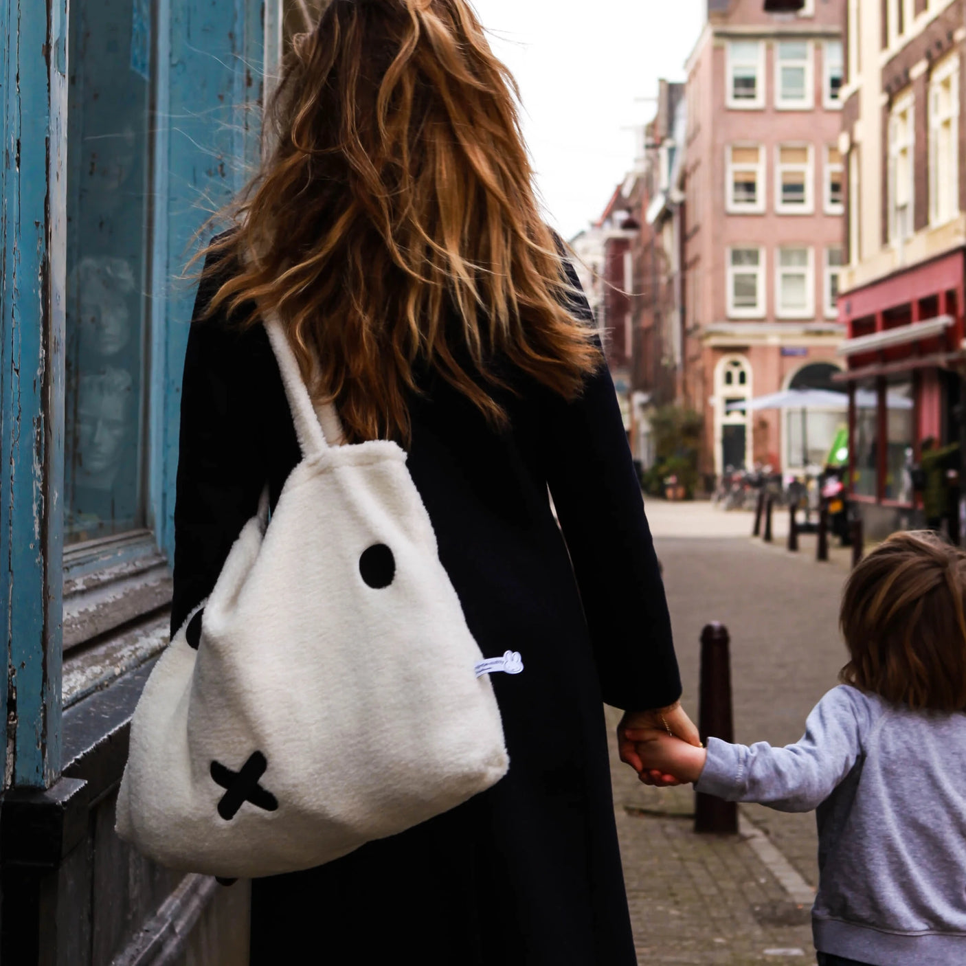 A woman with long hair and a black coat walks down the street, holding hands with a child. She proudly carries her sustainable fashion staple, the Miffy Shopping Bag from Miffy, which is crafted from recycled soft terry and showcases an adorable face design.